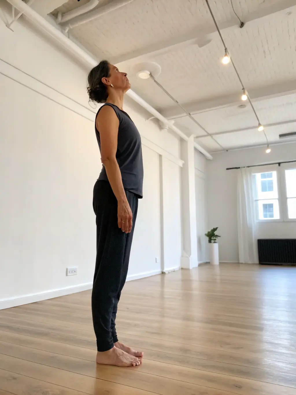 A photograph of a person in the Mountain Pose (Tadasana), demonstrating proper alignment and grounding, taken in a bright studio setting.